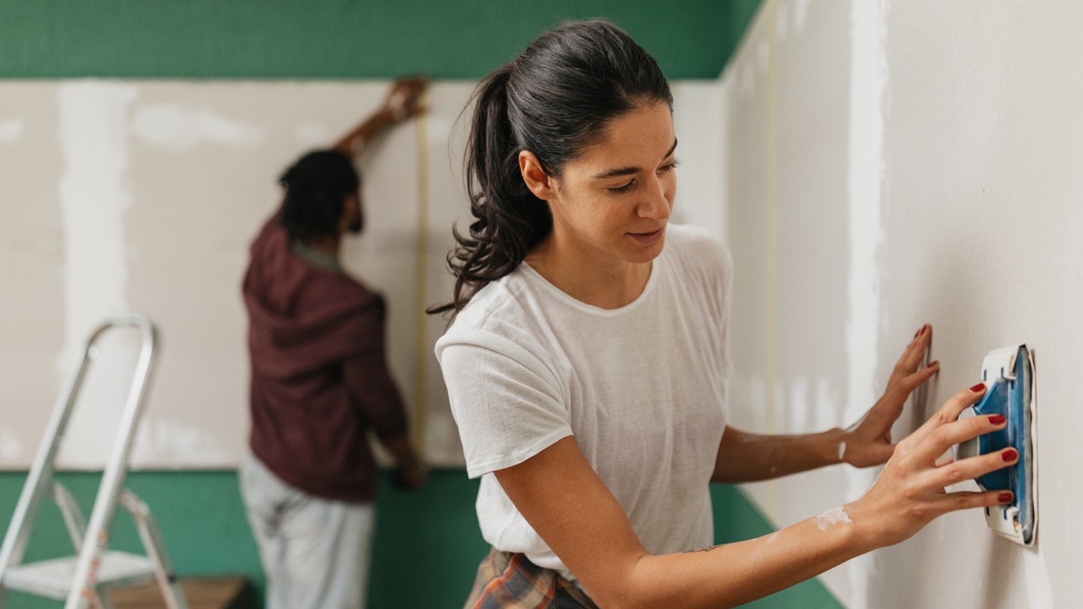 A woman sands a wall with a sanding block, while another person stands nearby and a ladder is visible in the background.