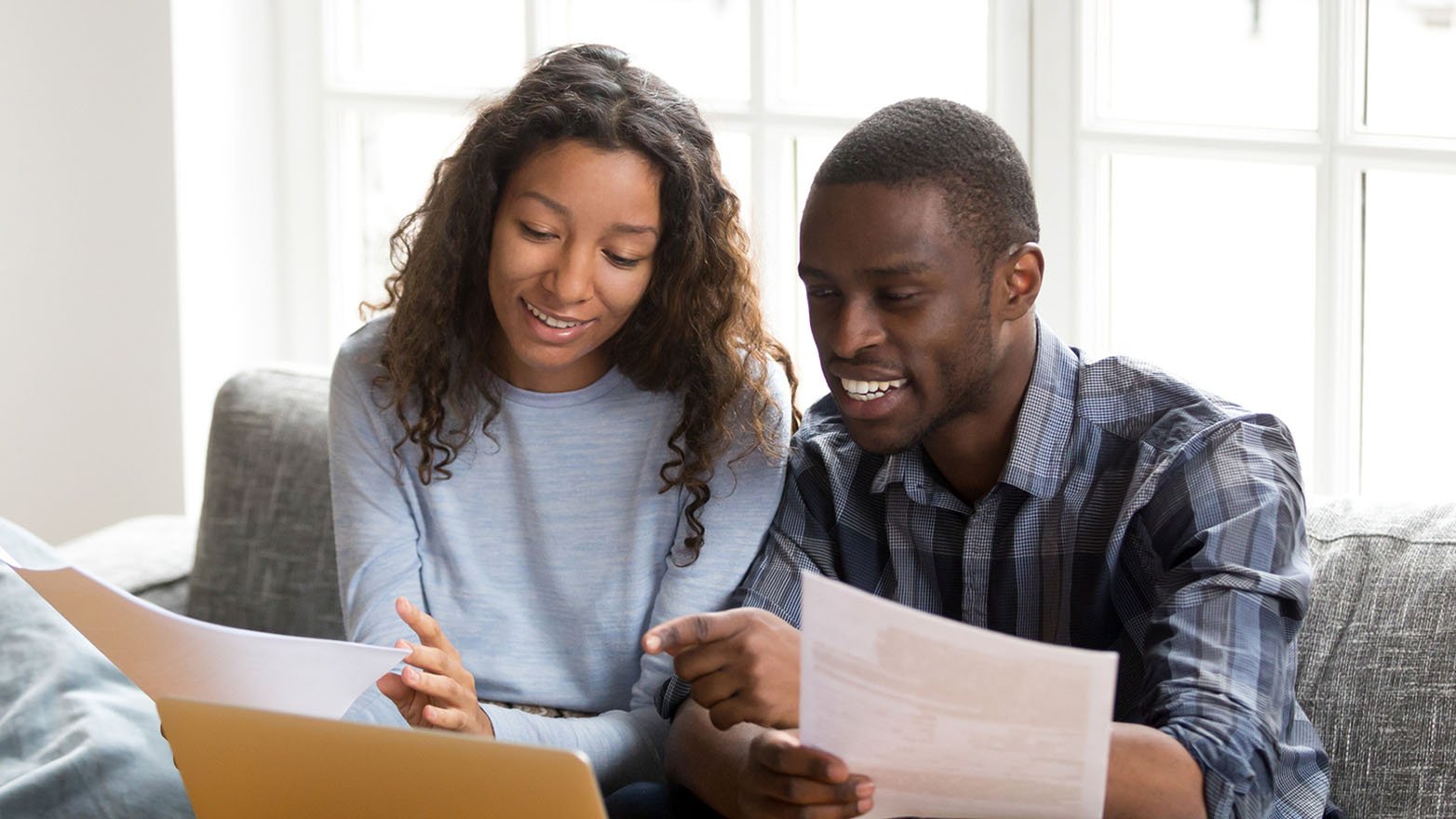 A young couple, a Black man and a biracial woman, are seated together on a grey sofa, reviewing documents and looking at a laptop. The man is pointing at a document he holds in his hand, and both are smiling, suggesting they are having a positive discussion.