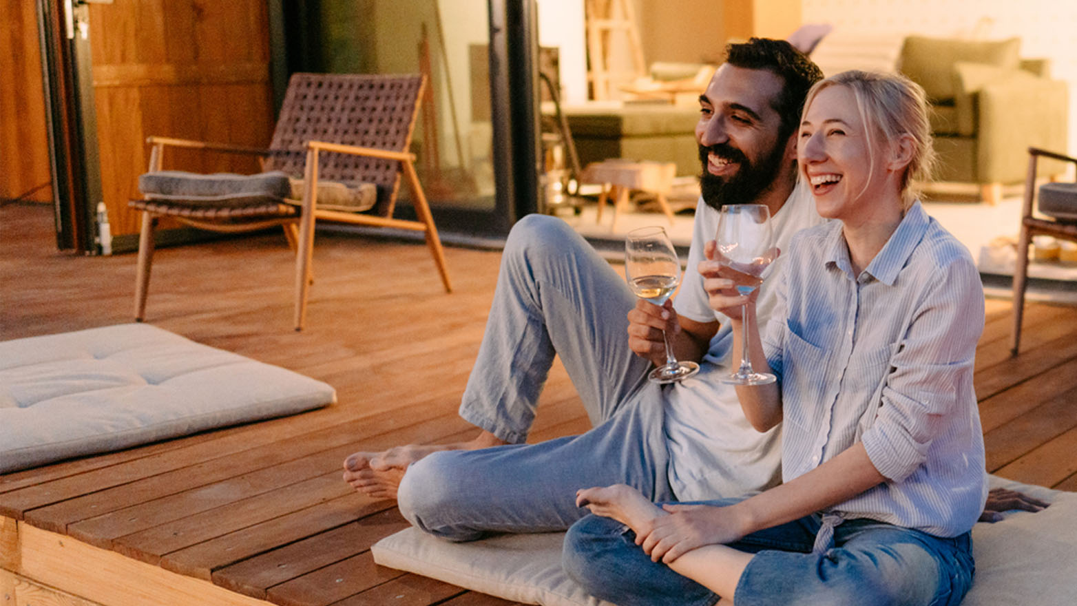 A bearded man and a blond woman are sitting on a wooden deck, holding wine glasses and laughing. They are barefoot and casually dressed. A cushioned mat sits beside them, and a wicker chair is visible in the background. Glass doors lead into the interior of the house.