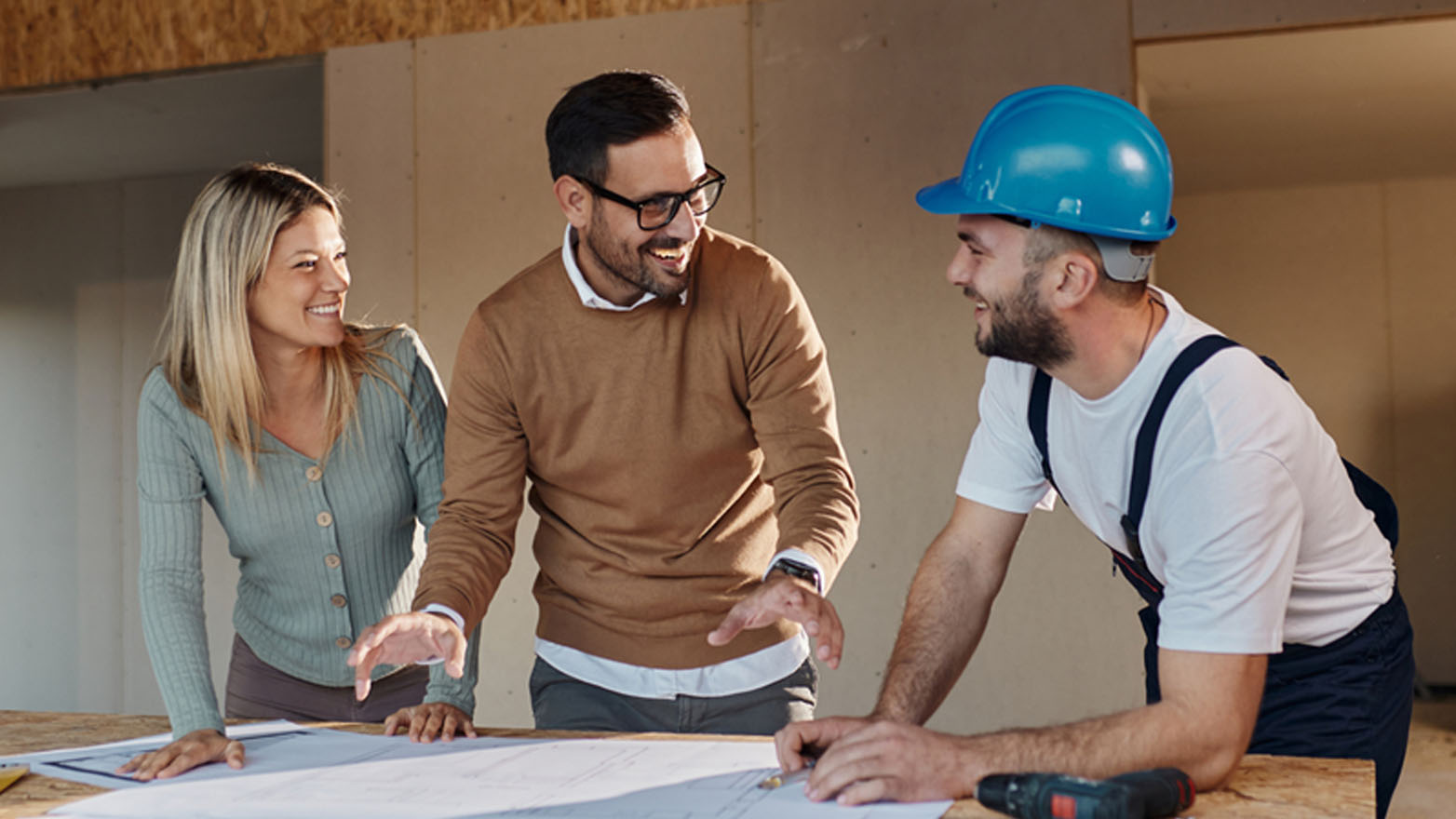 A smiling couple and a construction worker wearing a blue hard hat review blueprints on a wooden table in a partially constructed building. The couple looks engaged and pleased as the worker explains the plans, creating a sense of collaboration and excitement about the construction project.