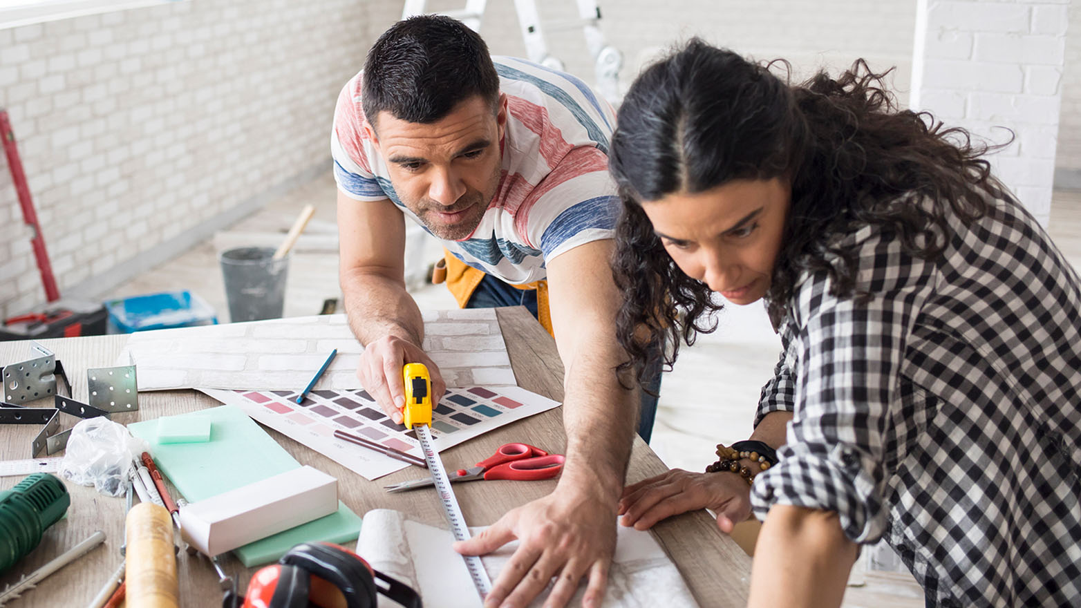 A couple is planning a home renovation. They are leaning over a table with a tape measure, paint swatches, and tools. They appear to be discussing the project, possibly measuring materials or selecting colors. The room behind them has a partially finished look, suggesting they are in the midst of the renovation process.