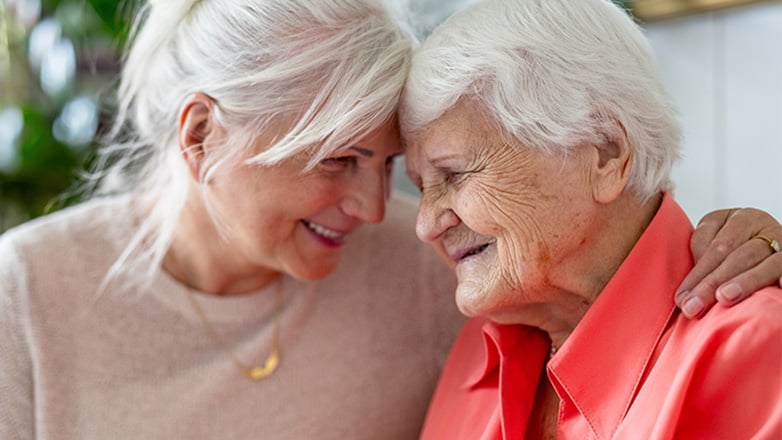 Two older women with white hair, likely friends or family, touch foreheads affectionately and share a warm smile.