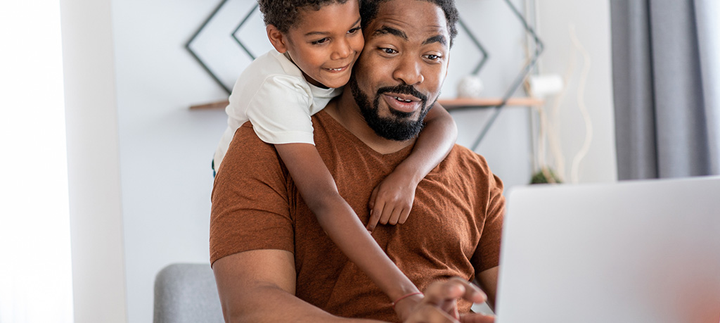 A medium shot of a Black man in a brown t-shirt working on a laptop with his son hugging him from behind. The father looks slightly surprised or amused at something on the screen. The son is smiling and looking in the same direction. The background is a simple, modern room with white walls and a geometric shelf. The father is seated on a grey chair.
