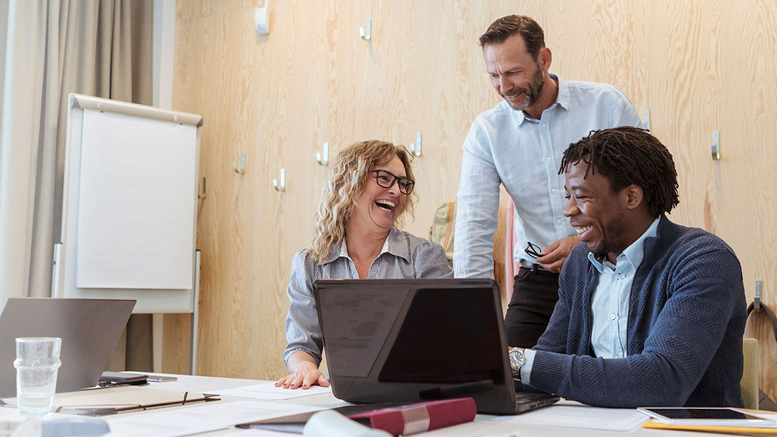 Three business colleagues, smiling, collaborate around a laptop.