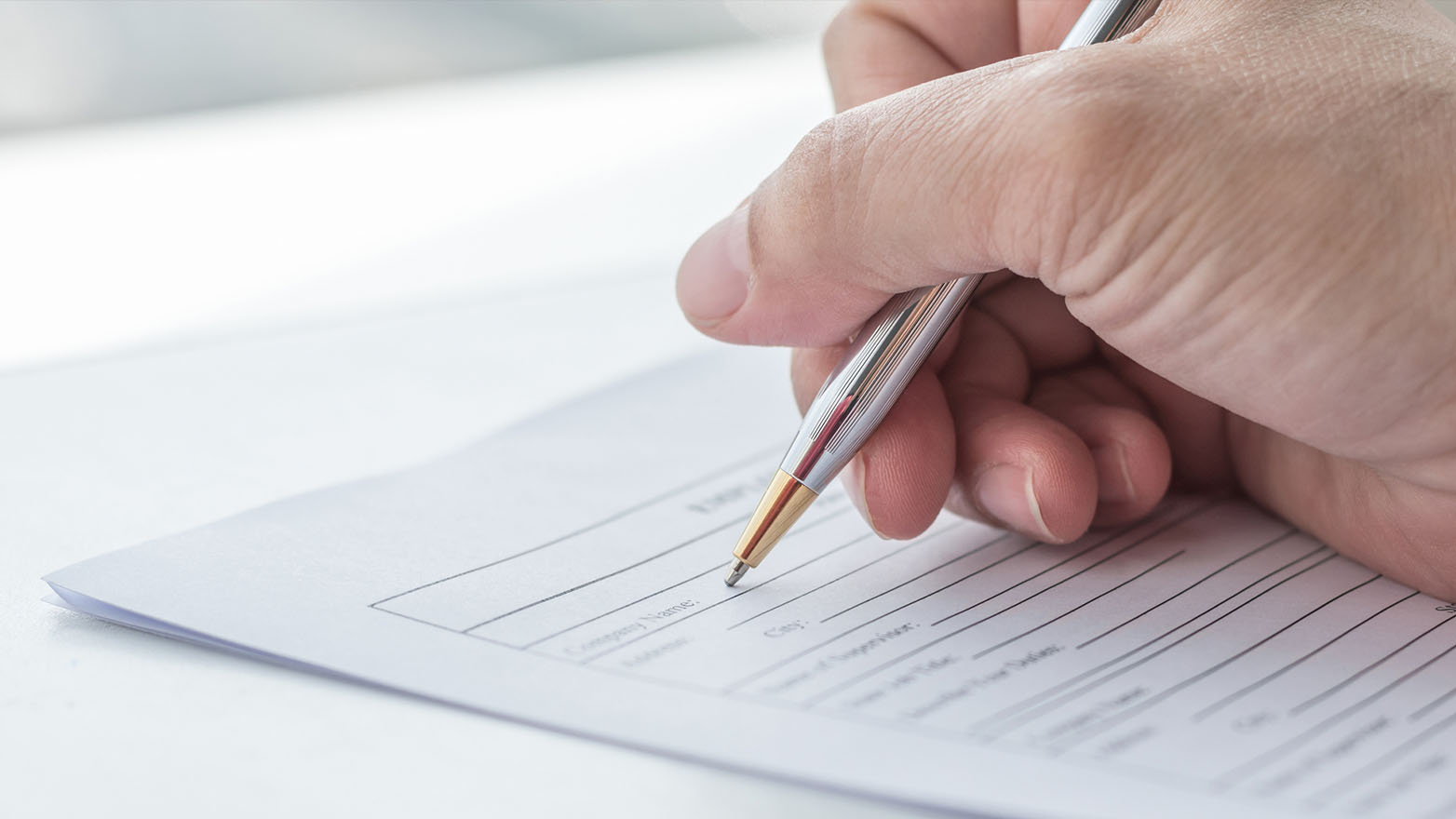 Close-up of a hand holding a silver pen, meticulously inscribing a form with spaces for company name, address, city, and the like.