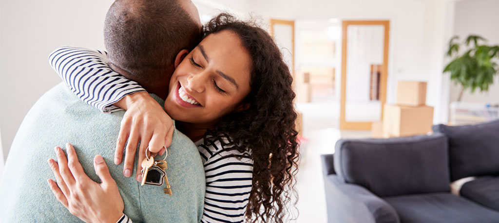 A jubilant woman with a cascade of curls embraces a bald gentleman, clutching the keys to their new abode. The scene unfolds in a luminous, capacious living space, with the remnants of a move—boxes and a somber sofa—in the backdrop.
