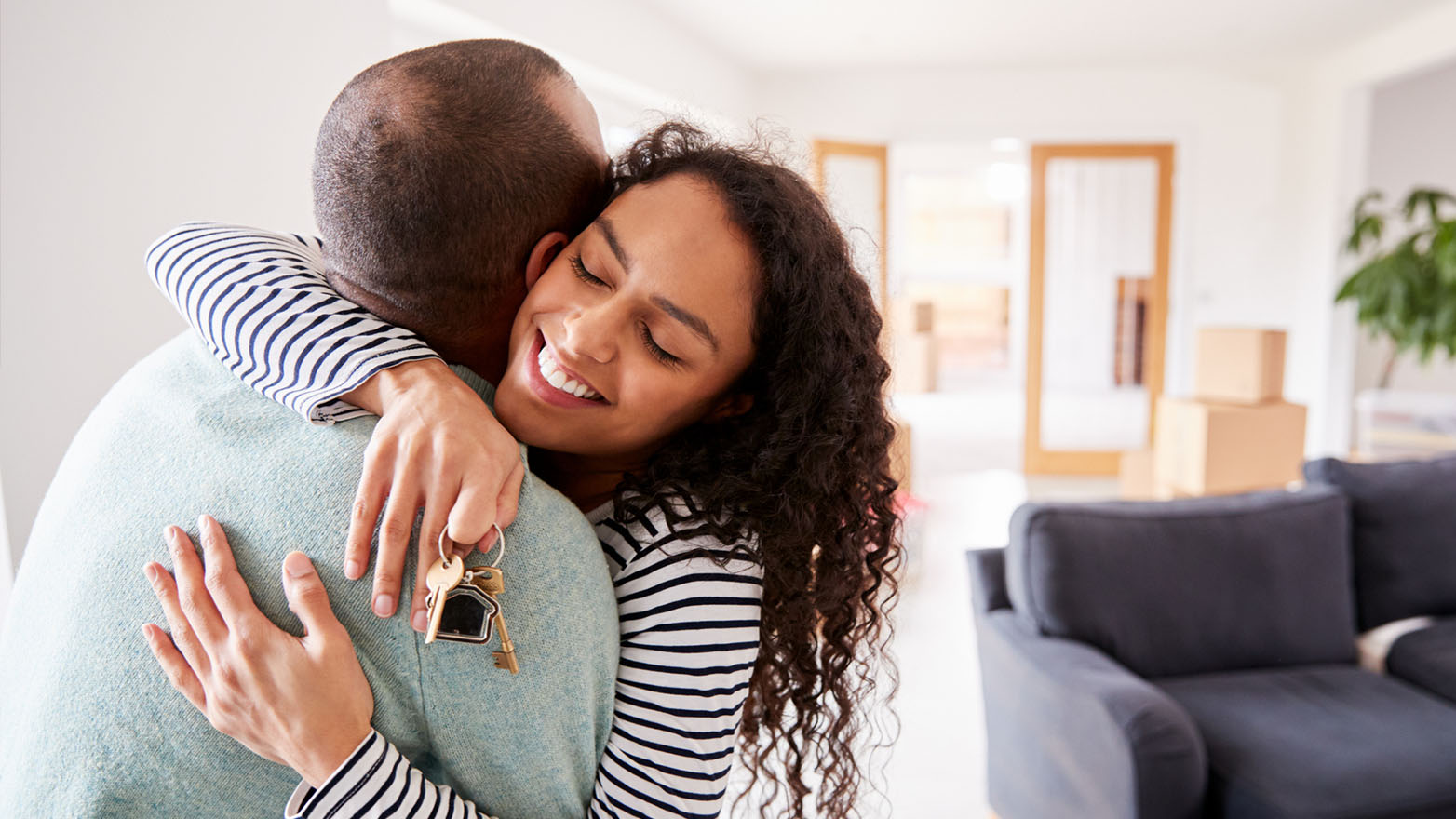 A jubilant woman with a cascade of curls embraces a bald gentleman, clutching the keys to their new abode. The scene unfolds in a luminous, capacious living space, with the remnants of a move—boxes and a somber sofa—in the backdrop.