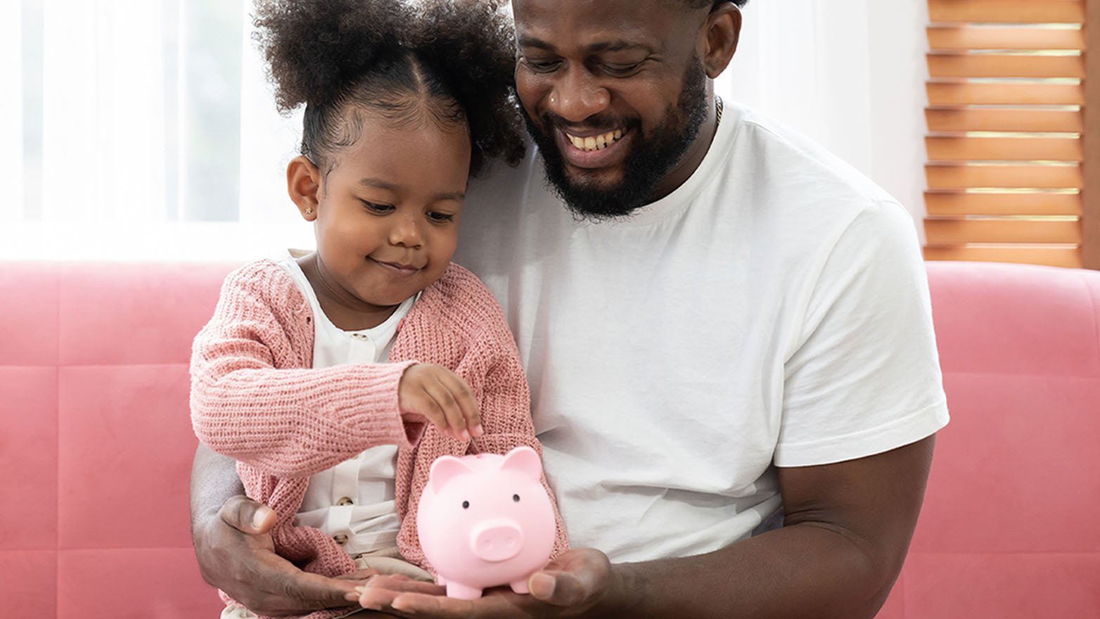 A smiling father gently holds his young daughter as she carefully deposits a coin into a traditional pink piggy bank.