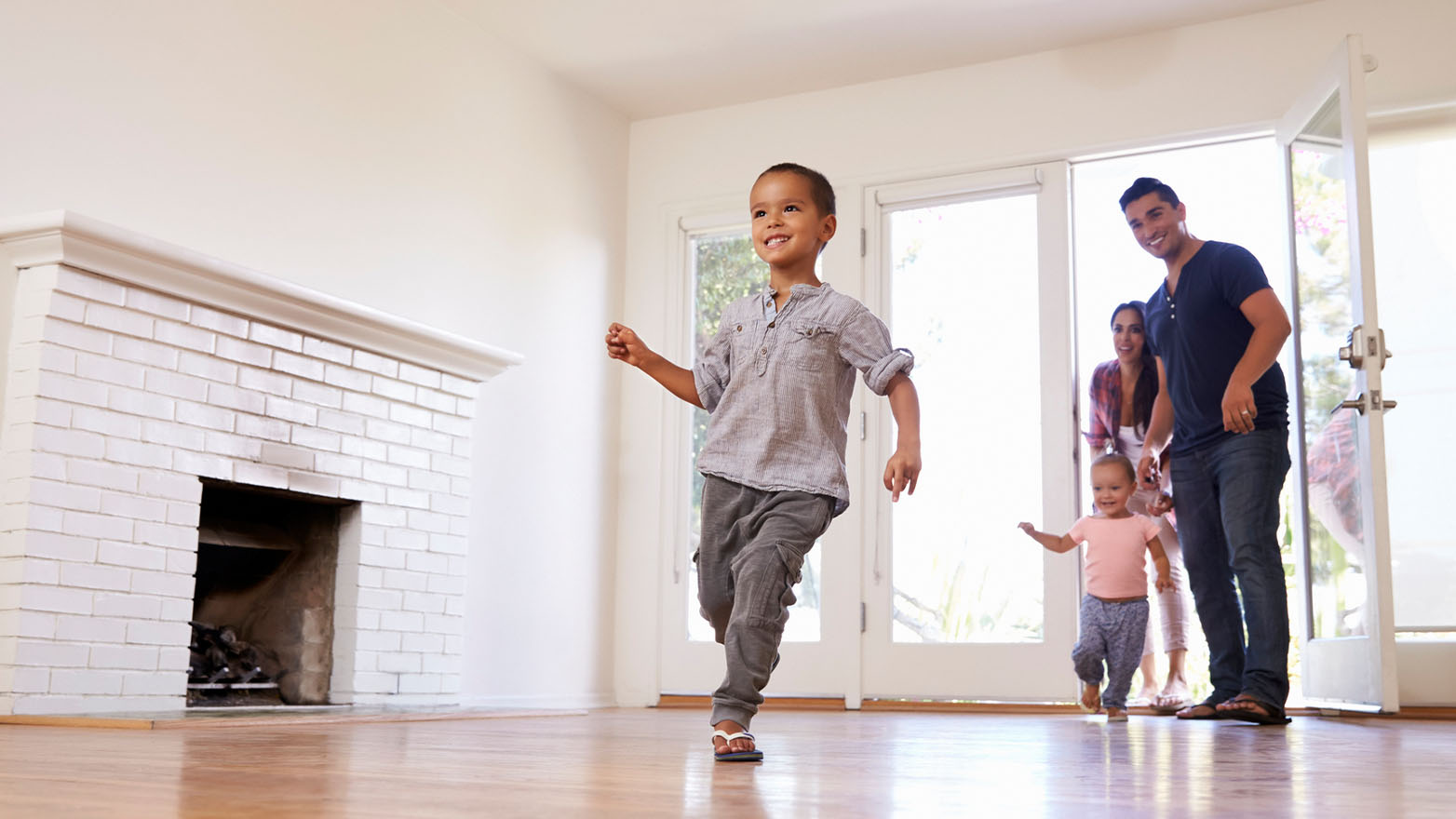 A family of four strolls in. Up front, a beaming lad makes a dash for the lens. In the back, a man and a woman beam too, with a wee one toddling along. There's a white brick fireplace, a window, and a glass door to the great outdoors. The floor? A fetching light brown wood.