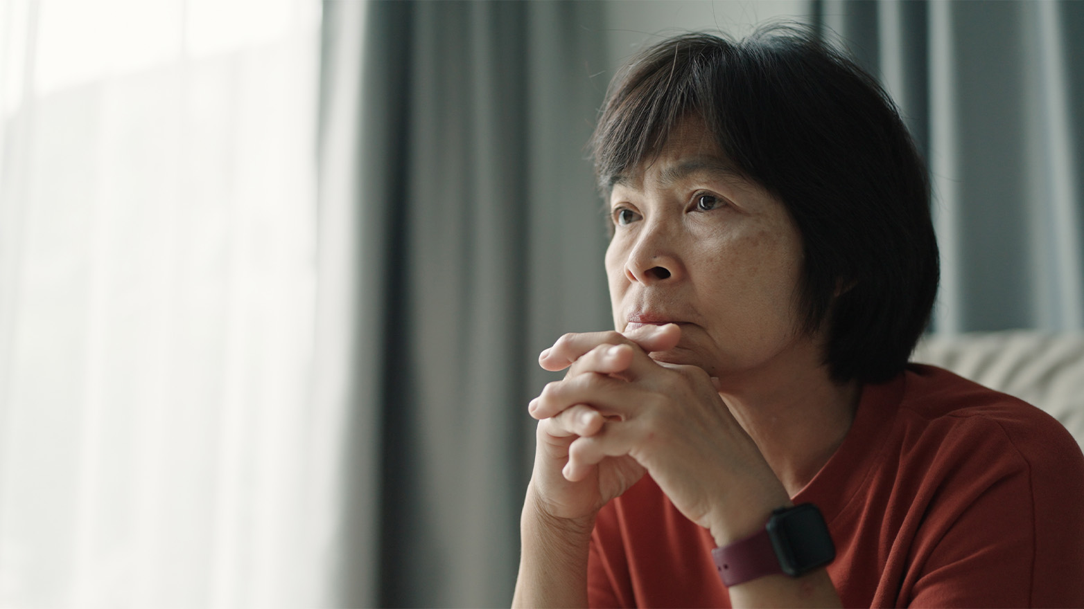 A woman with short, dark hair and Asian features sits in contemplative thought, her hands clasped under her chin as she gazes to the side.
