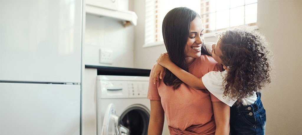 A medium shot of a young child hugging her mother in a laundry room. The mother is standing, facing left, with a laundry basket in front of her. She is wearing a short-sleeved pink t-shirt tied in a knot at the waist and denim shorts. The child has her arms wrapped around the mother's neck from the side. The girl is wearing a white top and jean shorts with buttons. Both are smiling at each other. In the background, there is an open washing machine, a refrigerator, and a window.