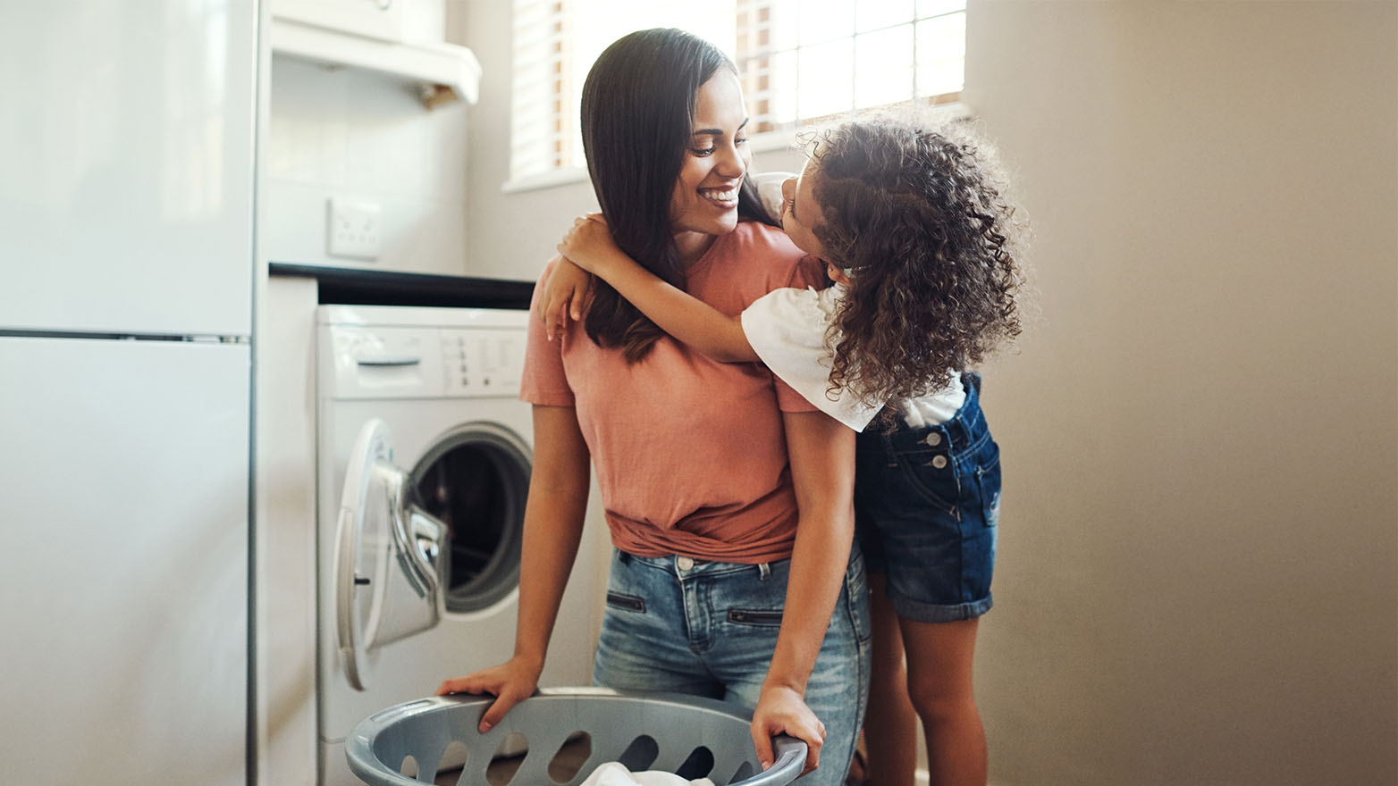 A medium shot of a young child hugging her mother in a laundry room. The mother is standing, facing left, with a laundry basket in front of her. She is wearing a short-sleeved pink t-shirt tied in a knot at the waist and denim shorts. The child has her arms wrapped around the mother's neck from the side. The girl is wearing a white top and jean shorts with buttons. Both are smiling at each other. In the background, there is an open washing machine, a refrigerator, and a window.