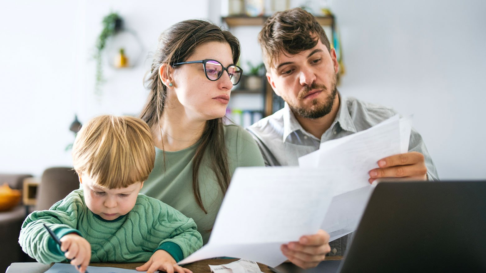 A young family, composed of a woman with glasses, a man with a beard, and a young boy, are seated at a table together. The man is holding several pieces of paper and displaying a concerned expression as he examines them. The woman is also looking in the same direction. The young boy is engaged in drawing on a piece of paper with a pencil. A laptop is visible in the foreground. The background reveals shelves and plants, indicating they are in a home environment.
