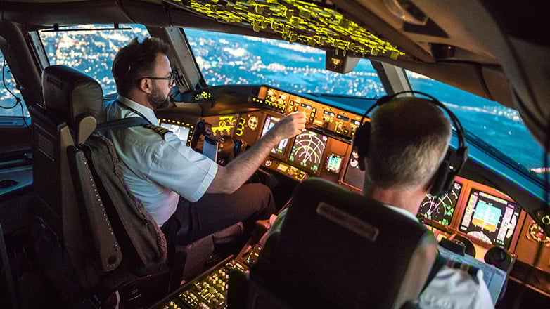 Two pilots in the airplane cockpit, one actively steering, fly over a cityscape at night, with numerous lights illuminating the landscape below.
