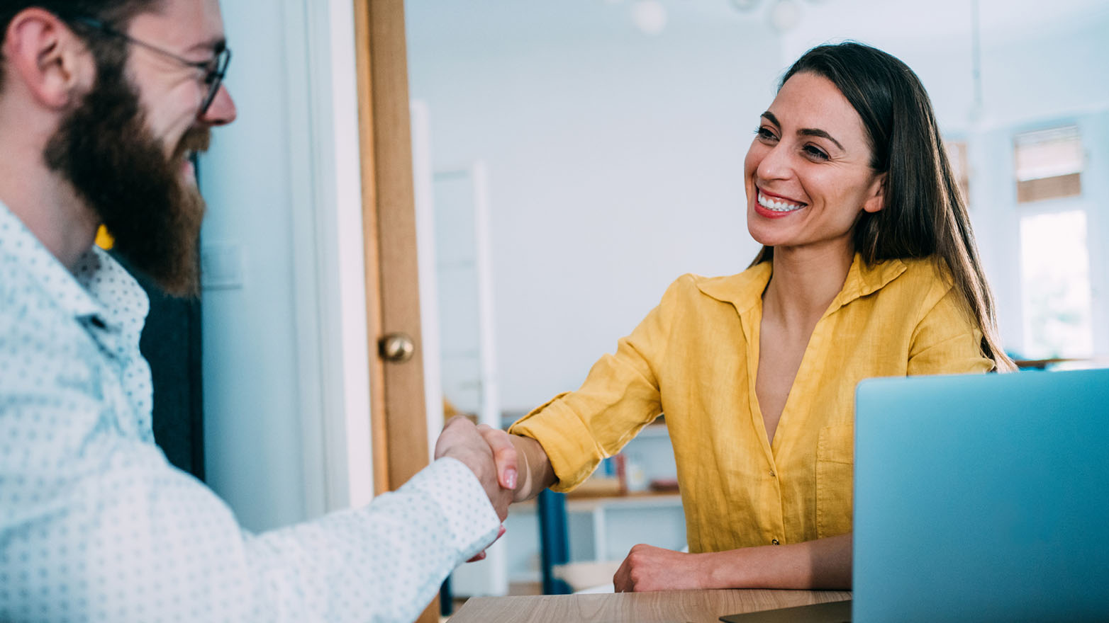 A smiling woman with long brown hair, donning a yellow blouse, and a man with a full beard in a light blue shirt with minuscule white dots are clasping hands in a well-lit office, signifying a meeting of minds, an accord, or a business deal. A laptop, partially obscured, is perched in front of the woman, hinting at a professional setting.
