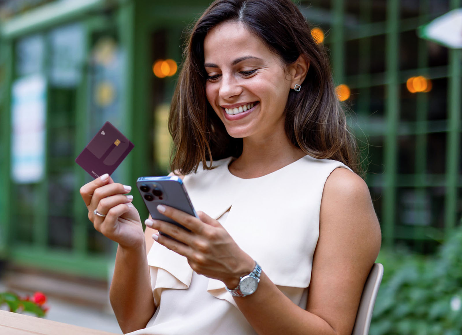 A happy young woman sits outside, smiling at her smartphone while holding up a dark purple credit card. She's wearing a cream-colored sleeveless blouse, a silver watch, and small flower-shaped earrings. A wedding ring adorns her left ring finger. The background features blurred greenery and a window with soft, warm lights reflecting on its surface. The overall impression is one of convenience and satisfaction with mobile banking or online shopping.