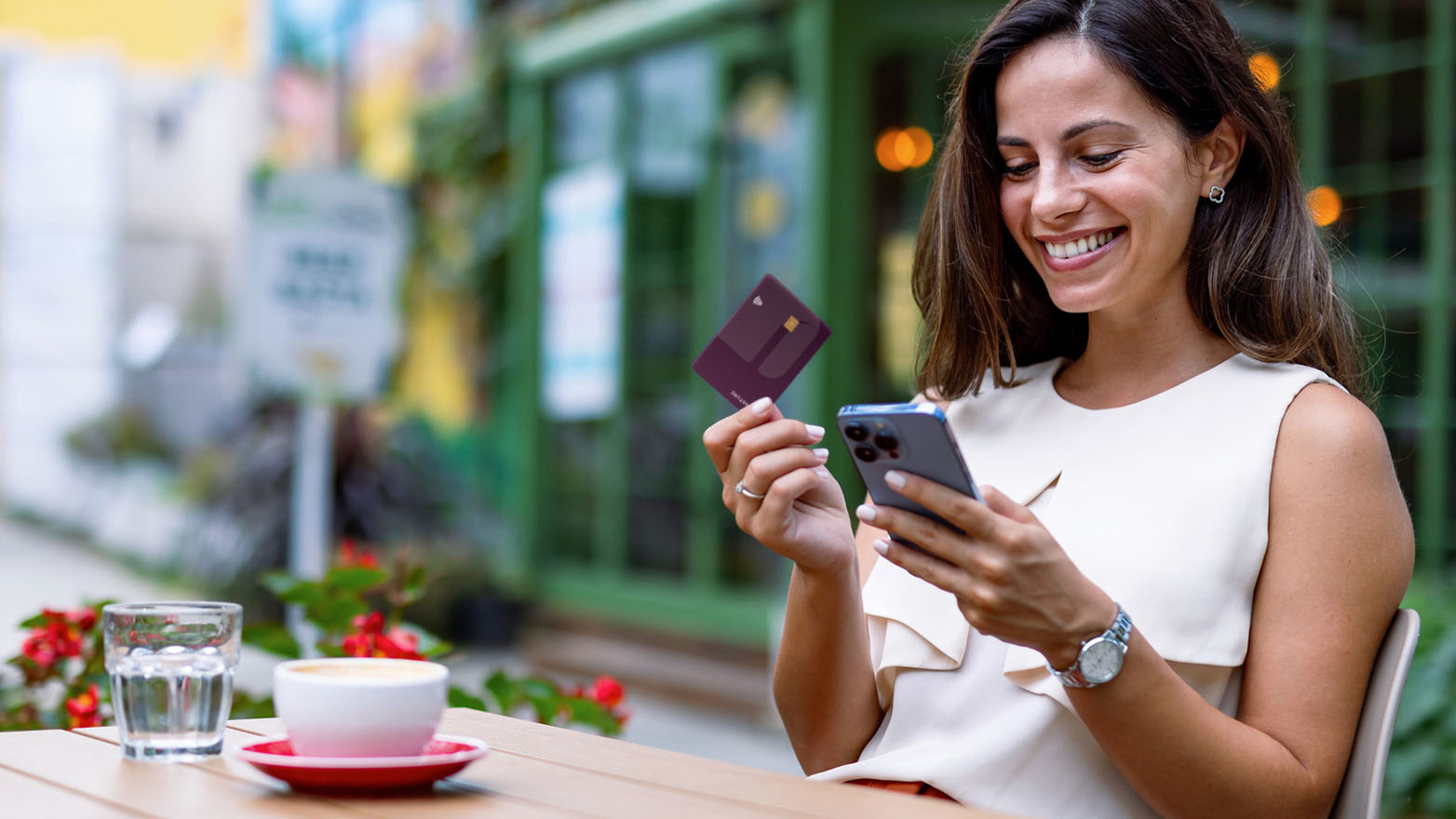 A happy young woman sits outside, smiling at her smartphone while holding up a dark purple credit card. She's wearing a cream-colored sleeveless blouse, a silver watch, and small flower-shaped earrings. A wedding ring adorns her left ring finger. The background features blurred greenery and a window with soft, warm lights reflecting on its surface. The overall impression is one of convenience and satisfaction with mobile banking or online shopping.