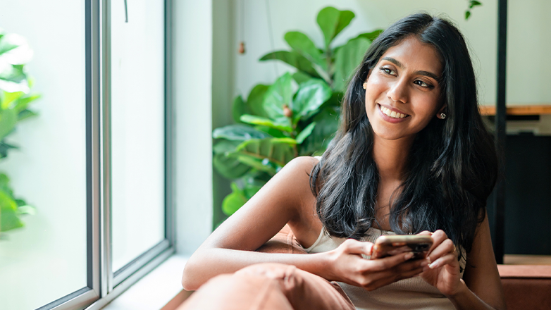 A young woman with long, dark hair sits on a couch, smiling as she looks to the side. She holds a smartphone in her hand, with a green plant and a window visible in the background.